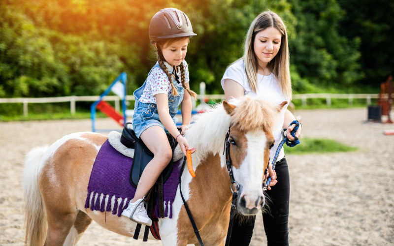 En flicka med ridhjälm rider på en ponny med hjälp av en vuxen.
