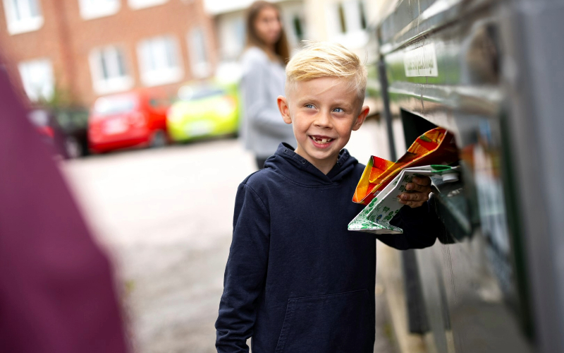En pojke med blont hår och glugg mellan framtänderna slänger pappersförpackningar på en återvinningsstation.