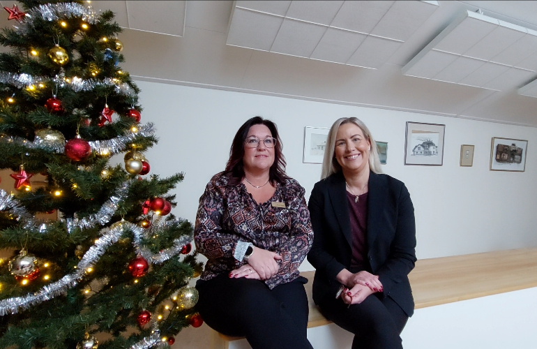 Anneli Mylly och Anna Nordqvist sitter på en bänk och ler mot kameran. Till deras högra sida står en julgran.