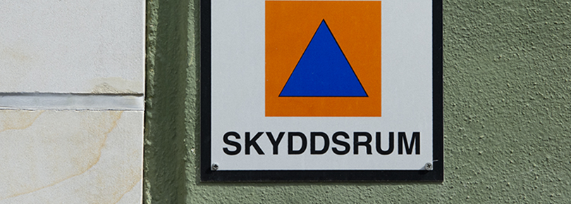 Skylt på en dörr till ett skyddsrum. Skylten består av en blå triangel mot en orange bakgrund.