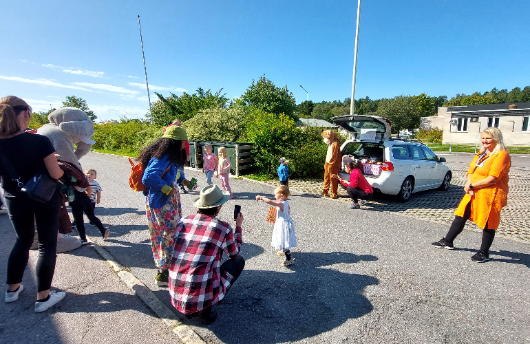 En person i en maskotdräkt tillsammans med en person i röd tröja delar ut godispåsar på parkeringen utanför Stora Vallahallen. Flera barn och föräldrar står runt bilen.